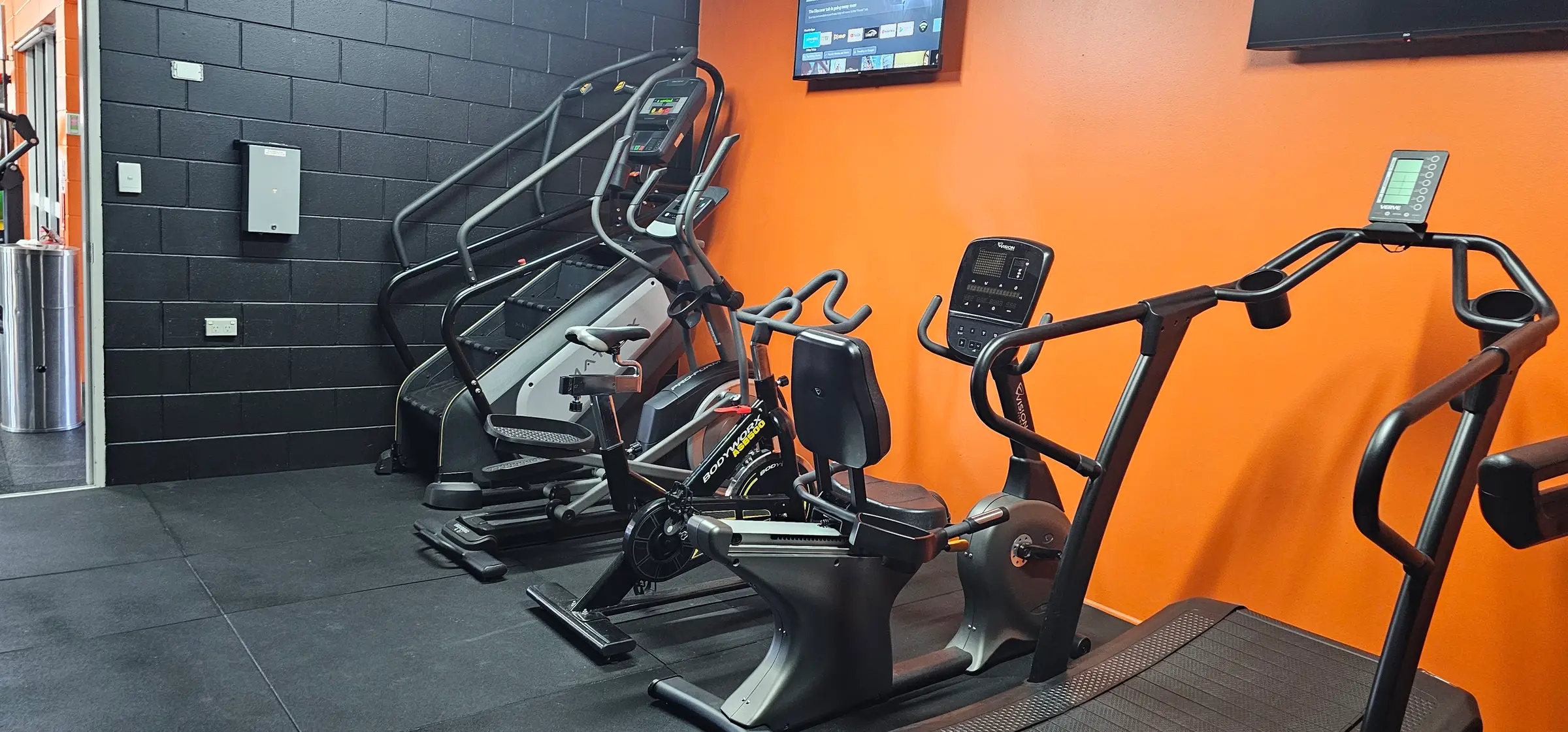 The cardio room at Get Fit Burdekin: a stairmill, elliptical and bikes against the orange and black walls, with a screen mounted above