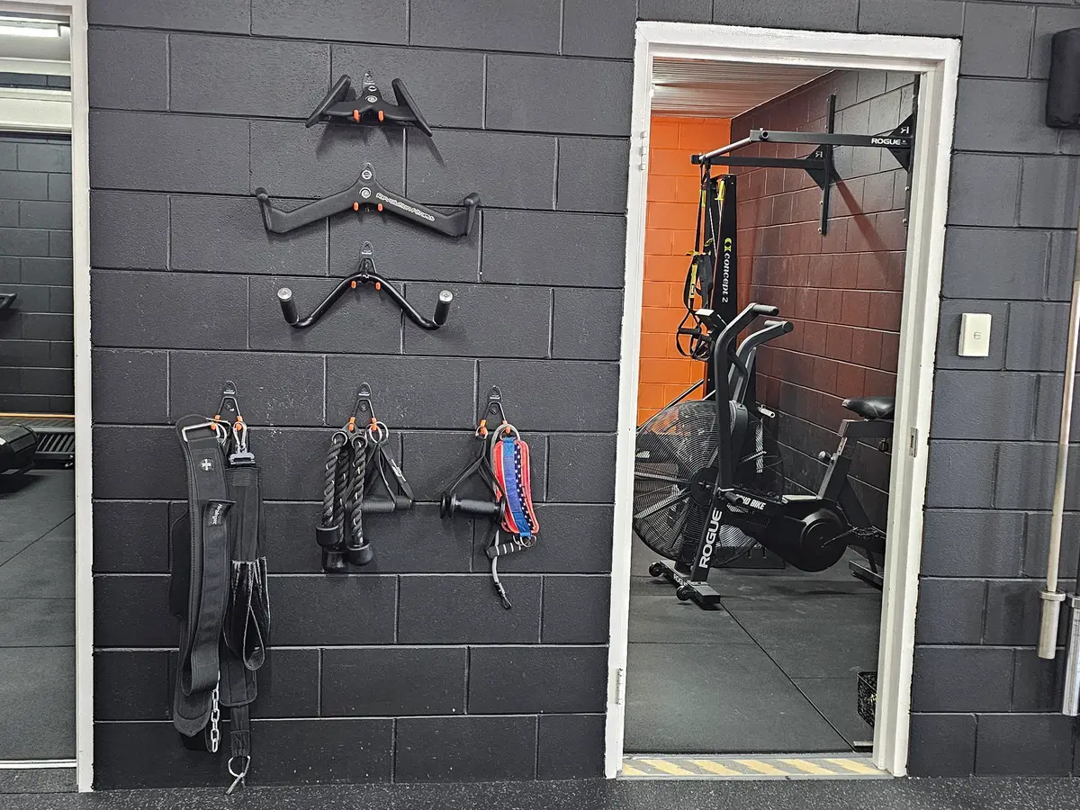 Cable handles, ropes and straps hung on the brick wall between two doorways, with barbells racked upright beside them