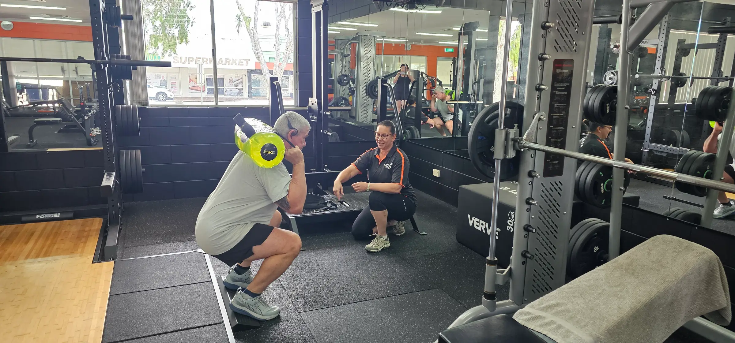 Yvette kneeling on the floor coaching a member through a squat with a yellow weight on his shoulder, beside the Smith machine at Get Fit Burdekin