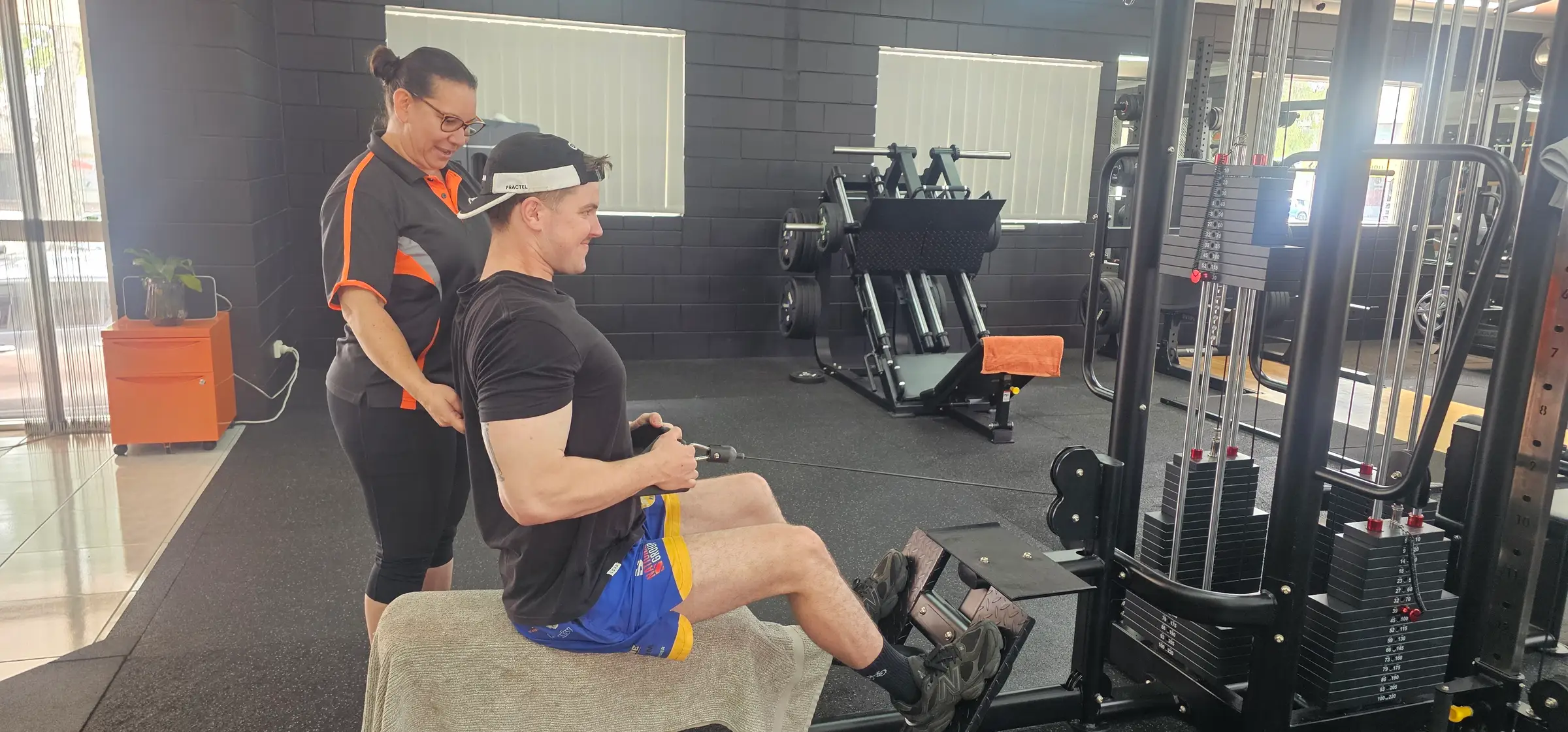 Yvette with a hand on a member's back, checking his posture on the seated cable row, with the leg press behind them at Get Fit Burdekin