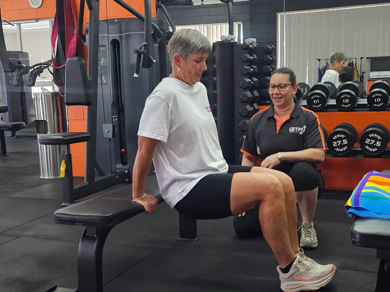 Yvette kneeling beside a member doing a split squat off a bench, watching the front knee, on the weights floor at Get Fit Burdekin